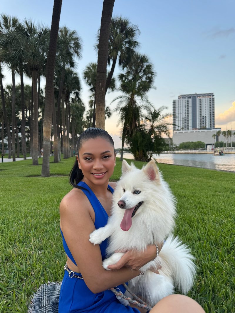 Devidasa Kawal in a royal blue dress, smiling and holding her white Pomeranian on a lawn by the water lined with palms