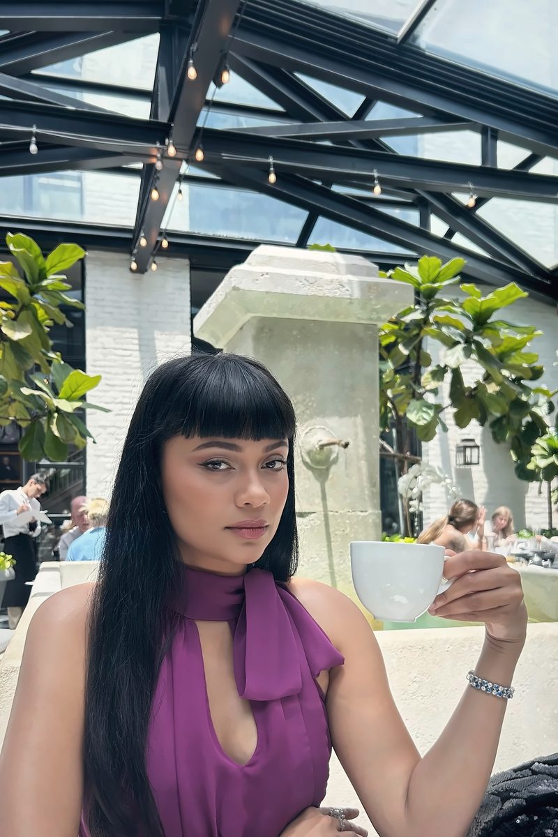 Devidasa Kawal in a purple halter top with a teacup, seated in a glass-roofed garden cafe