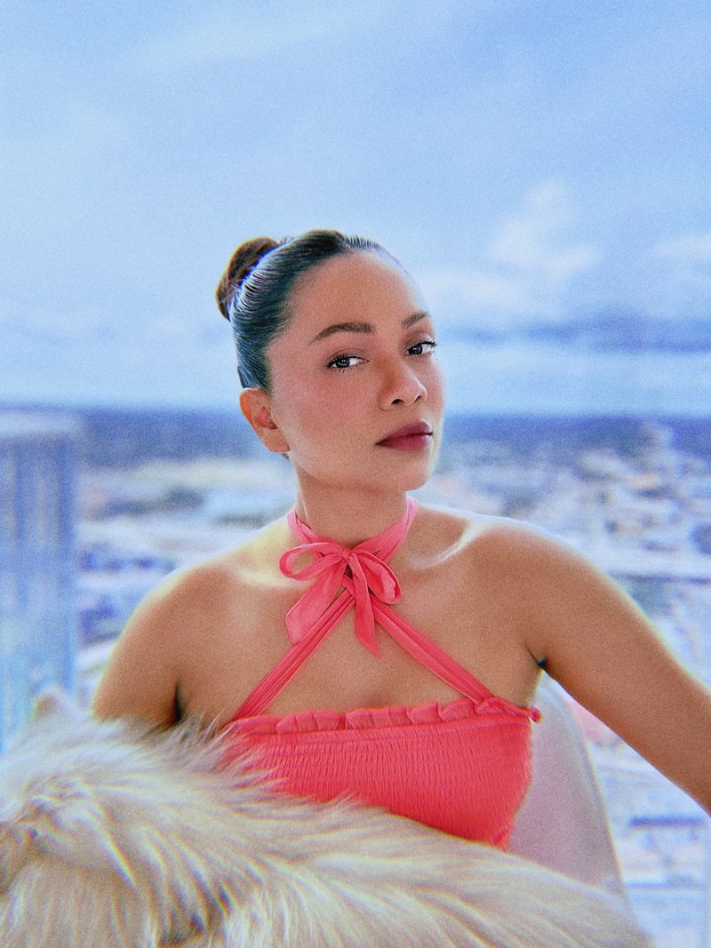 Portrait of Devidasa Kawal in a coral smocked halter top tied with a bow at the neck, hair in a sleek bun, with a city skyline behind her
