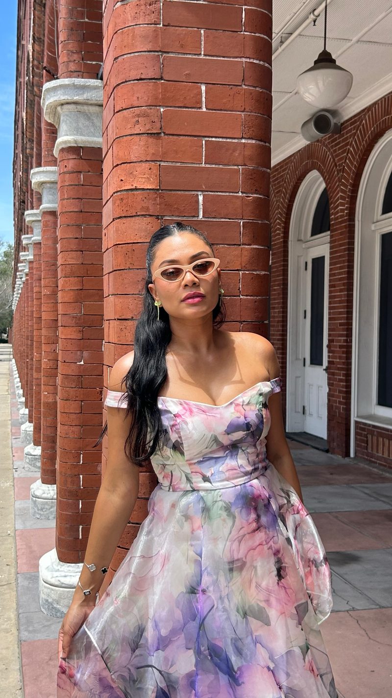 Devidasa Kawal in a floral organza off-the-shoulder gown and rose-tinted sunglasses, standing in a red brick colonnade
