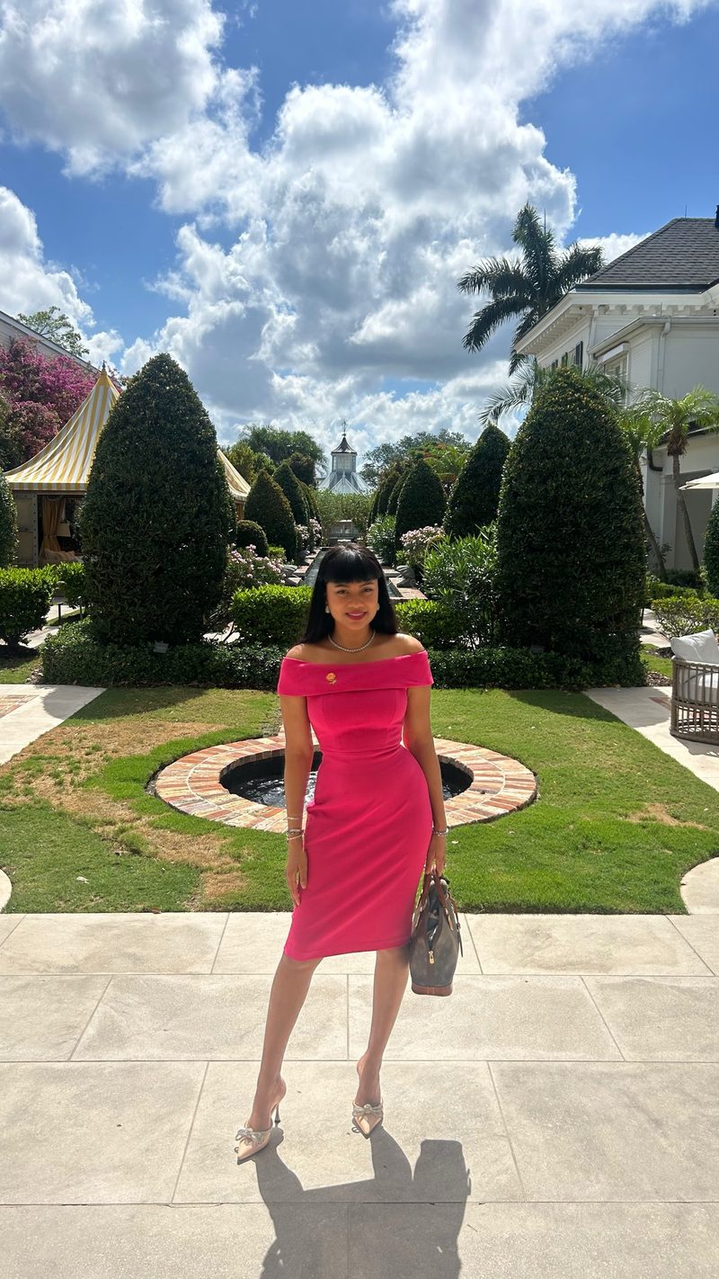 Devidasa Kawal in a fuchsia off-the-shoulder sheath dress, holding a small handbag in a manicured garden