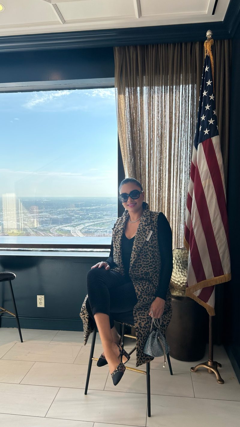 Devidasa Kawal in a long leopard-print vest and sunglasses, seated beside a high-rise window with a city view and an American flag