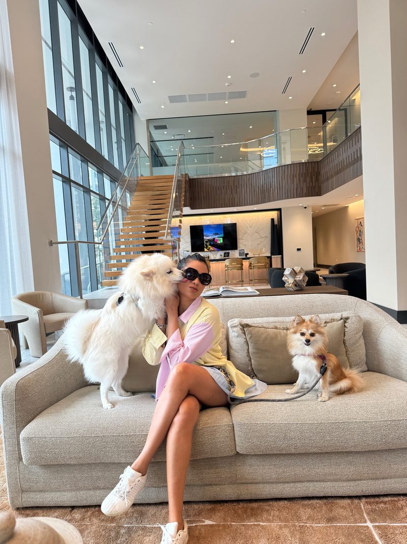 Devidasa Kawal in a pink and yellow shirt and sunglasses, seated on a sofa in a lobby with her two Pomeranians
