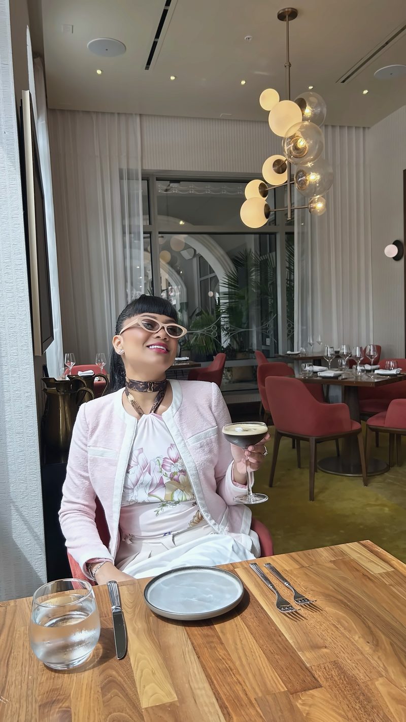 Devidasa Kawal in a pink tweed jacket and white sunglasses, raising a glass at a restaurant table