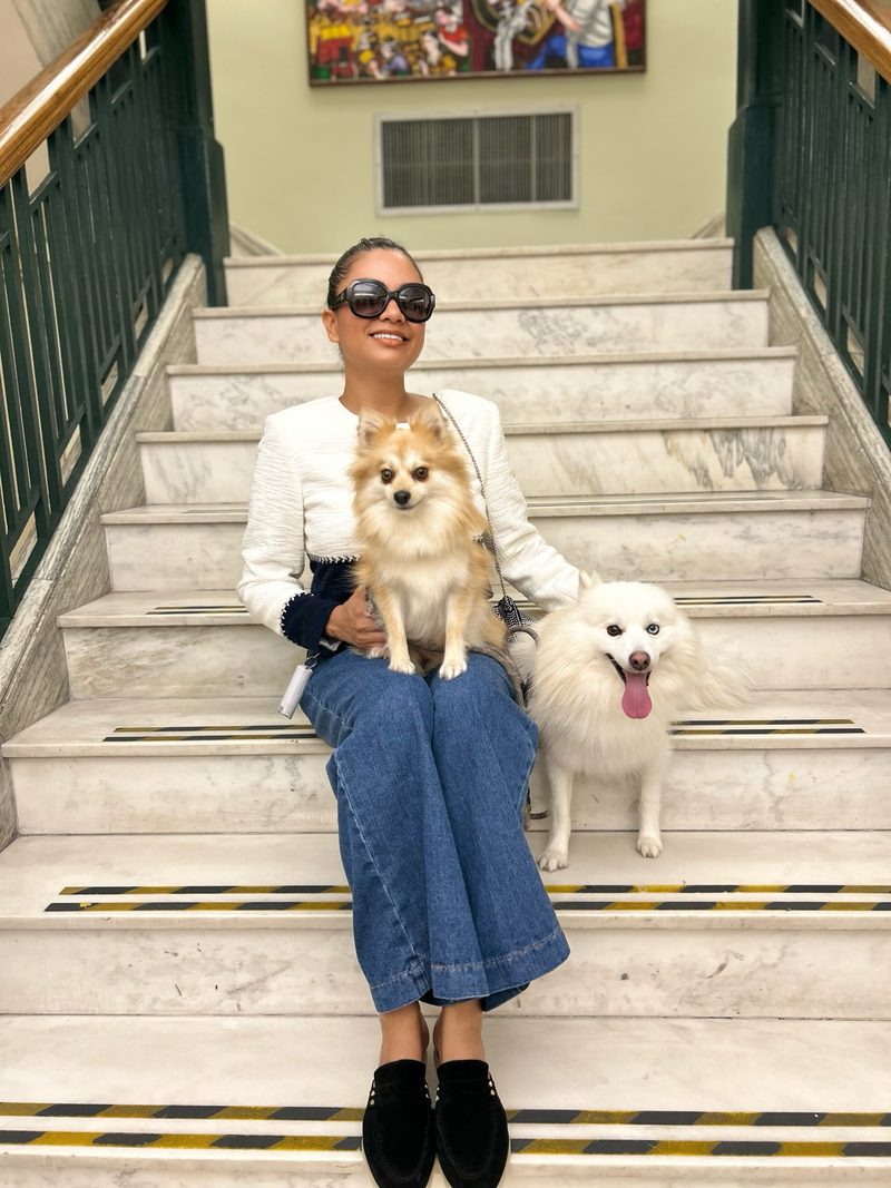 Devidasa Kawal in a white tweed jacket, wide-leg jeans and sunglasses, seated on marble stairs with her two Pomeranians