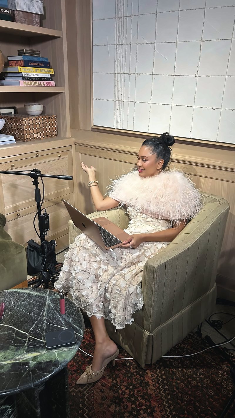 Devidasa Kawal in an ivory feathered dress, seated in a podcast studio with a laptop and microphone