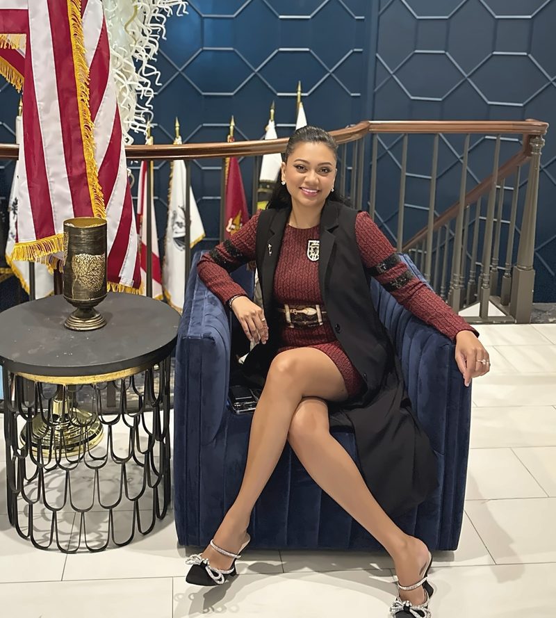 Devidasa Kawal smiling, seated in a navy velvet armchair in a burgundy dress and long black vest, with American and service flags against a navy geometric wall